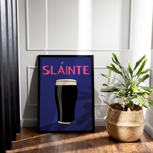 May include: Framed art print featuring a pint of dark stout with a creamy head, set against a deep blue background. The word "SL&Aacute;INTE" is displayed in pink. A woven basket with a green plant sits to the right.