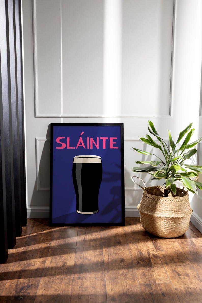 May include: Framed art print featuring a pint of dark stout with a creamy head, set against a deep blue background. The word "SL&Aacute;INTE" is displayed in pink. A woven basket with a green plant sits to the right.