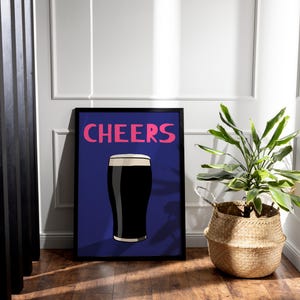 May include: A framed art print featuring a dark stout beer with a foamy head, set against a blue background. The word "CHEERS" is written in pink above the beer illustration. A potted plant in a woven basket sits to the right.