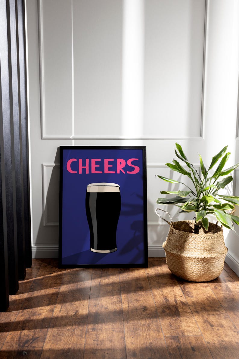 May include: A framed art print featuring a dark stout beer with a foamy head, set against a blue background. The word "CHEERS" is written in pink above the beer illustration. A potted plant in a woven basket sits to the right.