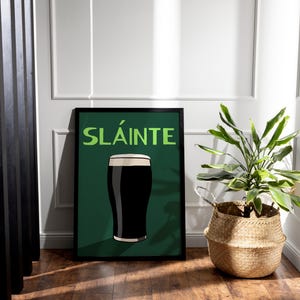 May include: Framed art print featuring a pint of dark stout with a creamy head, set against a dark green background. The word "SL&Aacute;INTE" is displayed in green. The print is in a black frame, leaning against a white wall. A woven basket with a plant is next to the print.