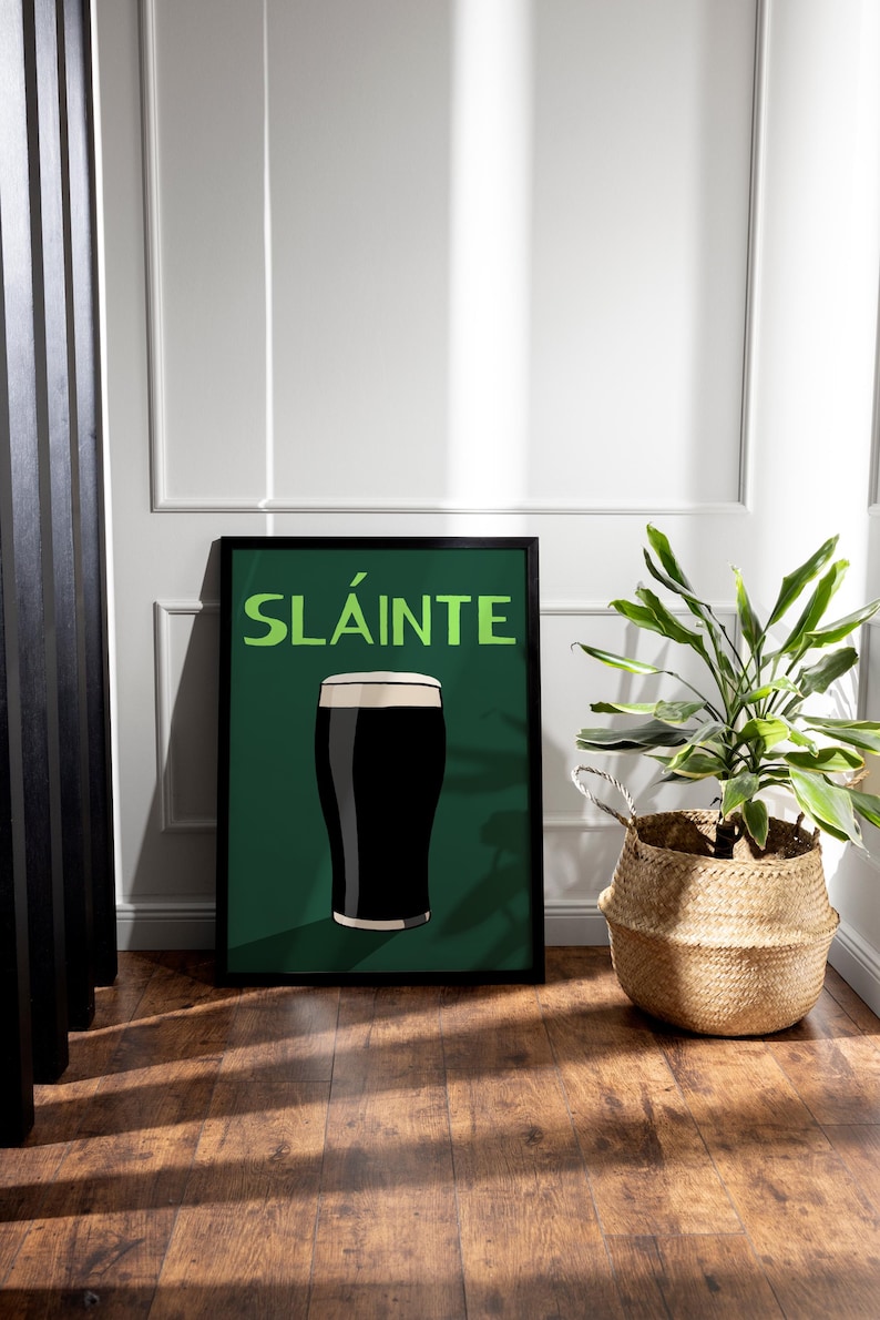 May include: Framed art print featuring a pint of dark stout with a creamy head, set against a dark green background. The word "SL&Aacute;INTE" is displayed in green. The print is in a black frame, leaning against a white wall. A woven basket with a plant is next to the print.