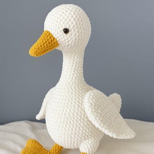 May include: A handmade white crochet goose plush toy with a yellow beak and feet. The goose has a long neck, small wings, and a textured, soft appearance. The toy is designed to stand upright and is likely intended for decorative or children's use.