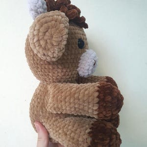 May include: A handmade crochet horse plushie in shades of brown, tan, and white. The plush toy has a fluffy texture, with a dark brown mane and hooves, and a white muzzle and ears. The horse is a soft, cuddly toy.