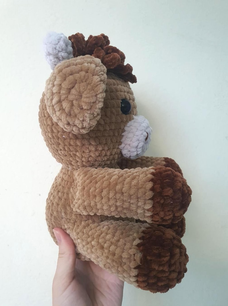 May include: A handmade crochet horse plushie in shades of brown, tan, and white. The plush toy has a fluffy texture, with a dark brown mane and hooves, and a white muzzle and ears. The horse is a soft, cuddly toy.