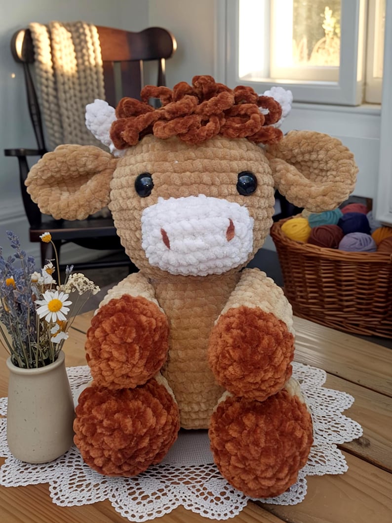 May include: A handmade, plush cow toy with a light brown body, white muzzle, and dark brown curly hair. The cow has large black eyes and is sitting on a wooden surface. The toy is made of soft, textured material and is a charming decorative item.