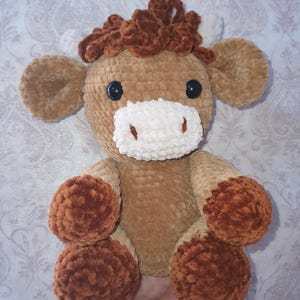 May include: A handmade, plush crochet cow toy in shades of brown and tan. The cow has a fluffy, textured body, with dark brown hooves and a matching tuft of hair. It features black safety eyes and a white muzzle.