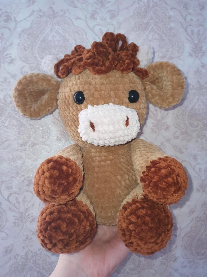 May include: A handmade, plush crochet cow toy in shades of brown and tan. The cow has a fluffy, textured body, with dark brown hooves and a matching tuft of hair. It features black safety eyes and a white muzzle.