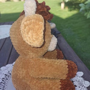 May include: A handmade plush toy in the shape of a deer, featuring a soft, textured fabric in shades of brown, tan, and cream. The deer has large ears, a fluffy mane, and a detailed face, perfect for a child's room or nursery.