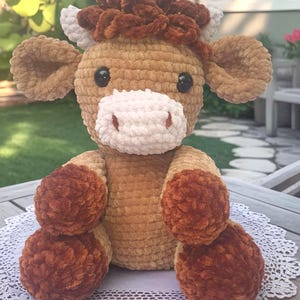 May include: A handmade crochet cow plushie with a light brown body, dark brown accents, and a white muzzle. The cow has black eyes and is sitting on a white lace doily. The plushie is a soft, cuddly toy.