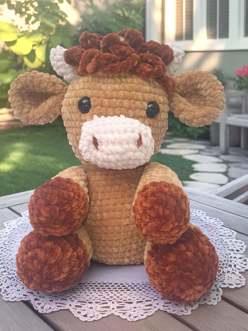 May include: A handmade crochet cow plushie with a light brown body, dark brown accents, and a white muzzle. The cow has black eyes and is sitting on a white lace doily. The plushie is a soft, cuddly toy.