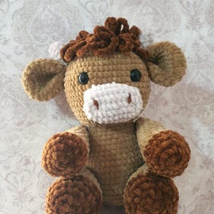 May include: A handmade crochet plush toy in the shape of a horse. The toy features a light brown body, dark brown mane and hooves, and a white muzzle. It has black button eyes and is designed to sit upright.