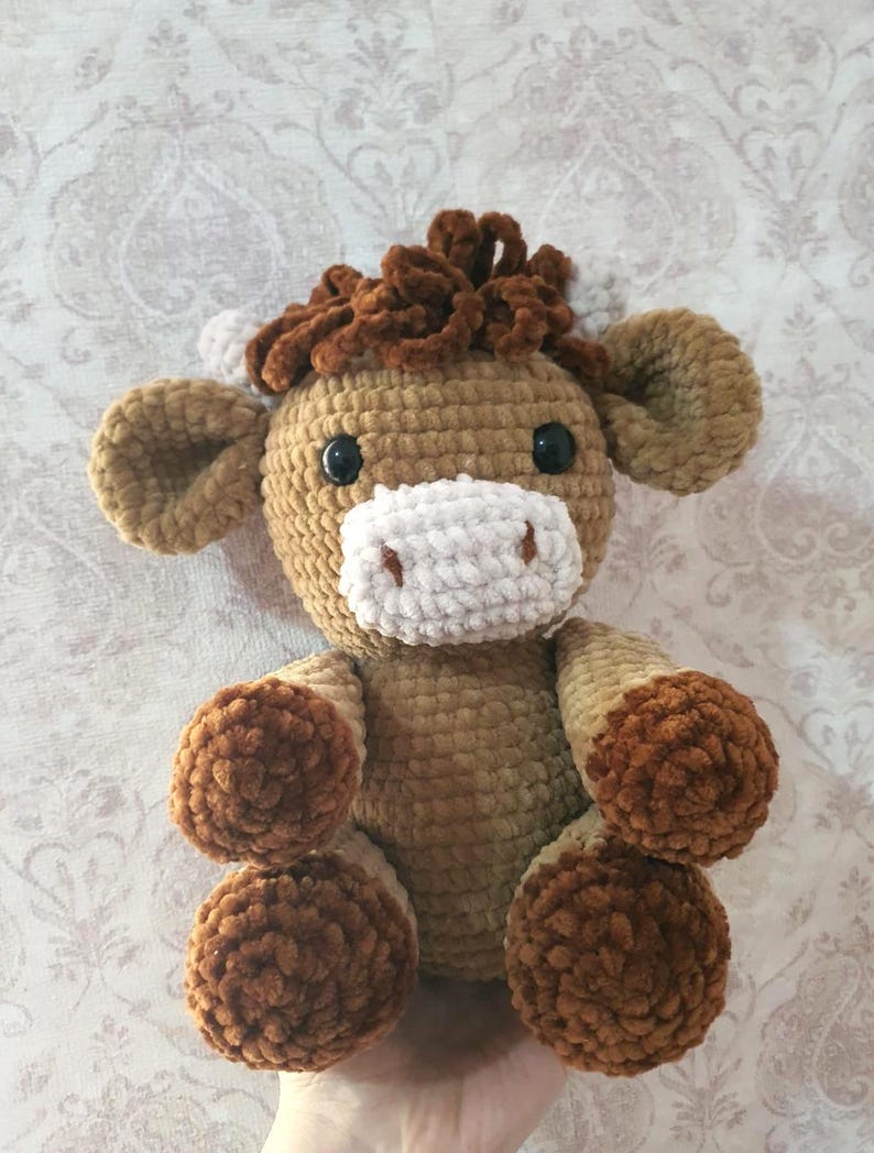 May include: A handmade crochet plush toy in the shape of a horse. The toy features a light brown body, dark brown mane and hooves, and a white muzzle. It has black button eyes and is designed to sit upright.