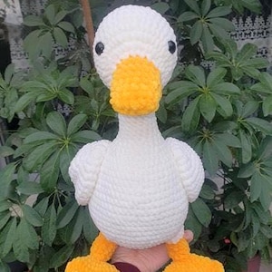 May include: A white crochet duck toy with a yellow beak and feet. The duck has black eyes and is held in front of green leaves. This soft toy is ideal for children. The duck is made of soft yarn.