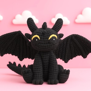 May include: A black crocheted dragon with yellow eyes and accents, wings spread, and a smiling expression. The dragon is set against a pink background with white cloud decorations. A handmade, whimsical item.