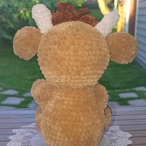 May include: A handmade, plush, brown crocheted deer toy. The deer has large, round ears, white antlers, and a brown mane. The toy is sitting on a white, lacy doily. The deer is made of soft, textured yarn.