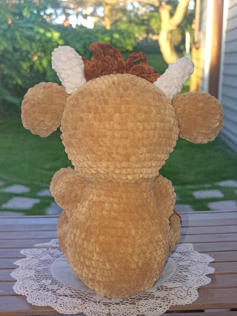 May include: A handmade, plush, brown crocheted deer toy. The deer has large, round ears, white antlers, and a brown mane. The toy is sitting on a white, lacy doily. The deer is made of soft, textured yarn.
