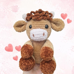 May include: A handmade, plush crochet cow in shades of brown, tan, and white. The cow has large, floppy ears, a white muzzle, and dark eyes. Its hooves and the hair on its head are a darker brown. The cow is sitting with its legs extended.