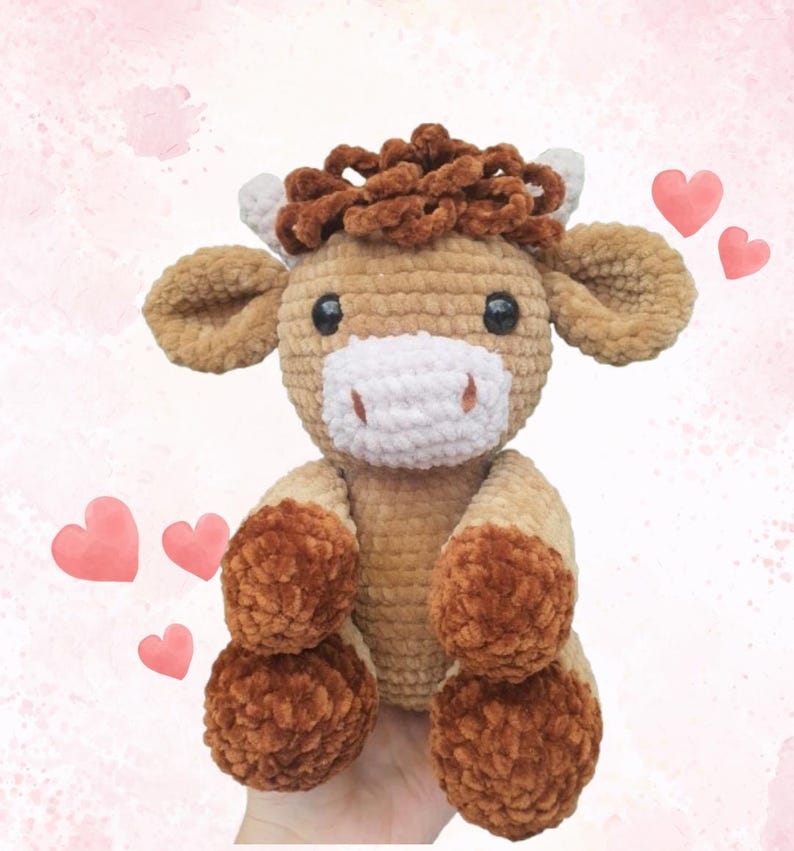 May include: A handmade, plush crochet cow in shades of brown, tan, and white. The cow has large, floppy ears, a white muzzle, and dark eyes. Its hooves and the hair on its head are a darker brown. The cow is sitting with its legs extended.