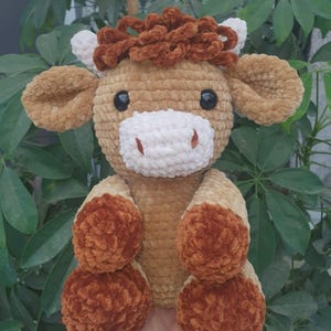May include: A handmade, plush cow toy in shades of brown, tan, and white. The cow has large, black eyes, a white muzzle, and fluffy brown accents on its head and feet. The toy is soft and cuddly, perfect for children.