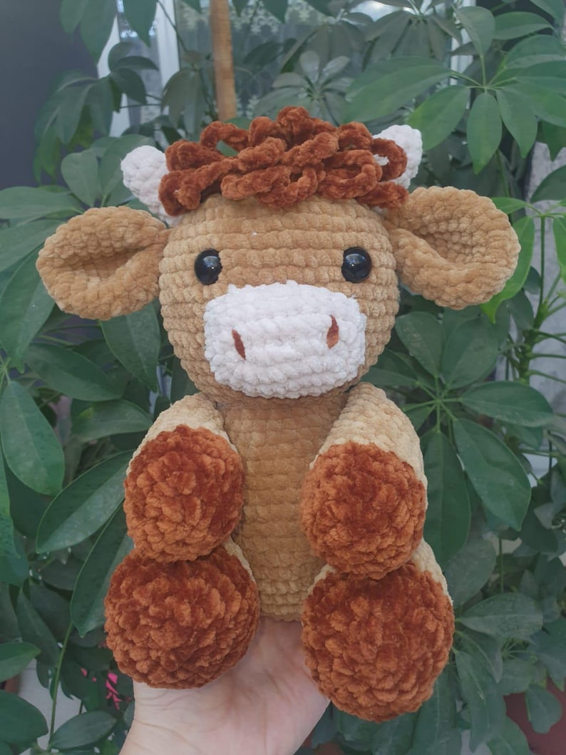 May include: A handmade, plush cow toy in shades of brown, tan, and white. The cow has large, black eyes, a white muzzle, and fluffy brown accents on its head and feet. The toy is soft and cuddly, perfect for children.