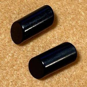 May include: Two glossy black cylindrical objects with rounded ends. The objects are lying on a textured, tan surface. The cylinders have a smooth, reflective surface, catching the light.