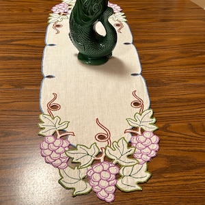 May include: A green ceramic fish-shaped pitcher sits on a white table runner with embroidered grape and leaf designs. The runner has a scalloped edge with blue and green trim. The table is made of wood.