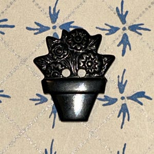 Op de afbeelding: Een zwarte metalen knoop in de vorm van een bloempot vol bloemen. De knoop heeft twee gaten om op te naaien en is geplaatst tegen een achtergrond met blauwe bloemmotieven.