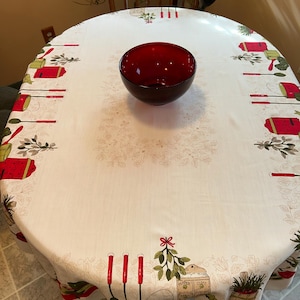 May include: A round, off-white tablecloth with a festive design featuring red and green gardening tools, gift boxes, and floral accents. A dark red glass bowl sits in the center. The tablecloth is on a round table.