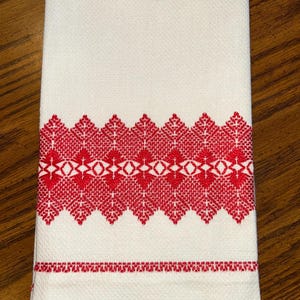 May include: White kitchen towel with a red embroidered floral design. The towel features a detailed pattern of stylized leaves and geometric shapes, adding a decorative touch to the kitchen.