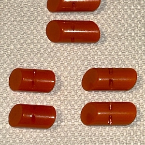 Puede incluir: Seis botones cilíndricos de color marrón rojizo con un extremo ligeramente angulado. Cada botón tiene una pequeña ranura horizontal. Los botones están dispuestos sobre una superficie texturizada de color gris claro.