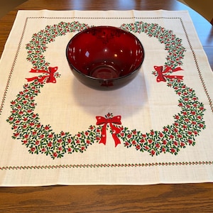 May include: A red glass bowl sits on a white tablecloth with a Christmas wreath design. The wreath is made of green leaves and red berries, with red bows. The tablecloth is on a wooden table.