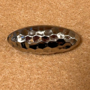 May include: A silver-colored, oval-shaped object with a textured, honeycomb-like surface. The object is reflective, catching light and creating highlights. The item appears to be a decorative element or jewelry component.