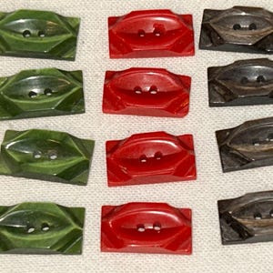 May include: A collection of rectangular buttons in various colors. The buttons are in shades of green, red, and dark brown. Each button has two holes for sewing and a raised, geometric design. The buttons are likely for clothing or craft projects.