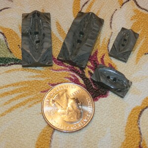 May include: A collection of four gray rectangular buttons with a carved leaf design and two holes for sewing. A U.S. quarter coin is next to the buttons for size comparison.