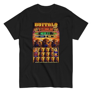 Puede incluir: Camiseta negra con un gráfico colorido de una máquina tragamonedas con el texto "BUFFALO LINK". El diseño incluye imágenes de búfalos y símbolos de cartas. También se puede ver el texto "YOU DON'T CHASE THE BUFFALO, THE BUFFALO CHASES YOU".