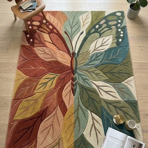 May include: A rectangular rug with a butterfly design. The rug features a central butterfly shape with leaf-like wings in shades of brown, orange, yellow, green, and blue. The rug is placed on a light wood floor.