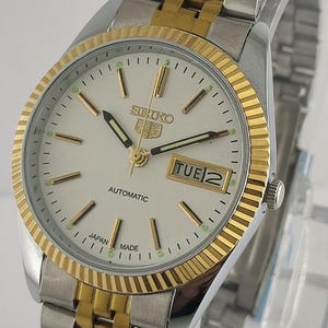 Pode incluir: Um relógio de pulso automático Seiko com pulseira e caixa em aço inoxidável prateado e dourado. O mostrador do relógio é branco com marcadores e ponteiros dourados e um visor de dia e data. O relógio é marcado "Japan Made".