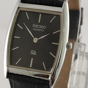 Könnte beinhalten: Eine rechteckige Seiko Quartz-Uhr mit schwarzem Zifferblatt und silberfarbenem Gehäuse. Die Uhr hat ein schwarzes Lederarmband und silberne Zeiger. Auf dem Zifferblatt sind das Seiko-Logo und das Wort "Quartz" zu sehen.