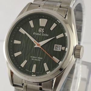 May include: A silver-toned stainless steel wristwatch with a dark green textured dial. The watch features a Grand Seiko logo, a date window displaying 29, and text reading HI-BEAT 36000 80 HOURS. It has a metallic link bracelet and a crown on the side.