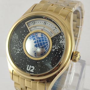 May include: Gold-toned wristwatch with a black face featuring a white and blue globe design. The watch has a gold band and a crown with a blue stone. The watch face also displays the text "U2".