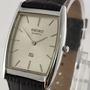 Könnte beinhalten: Rechteckige Seiko Quartz-Uhr mit silberfarbenem Zifferblatt und schwarzem Lederarmband. Das Zifferblatt zeigt das Seiko-Logo und ein kleines Design. Das Uhrengehäuse ist silberfarben.