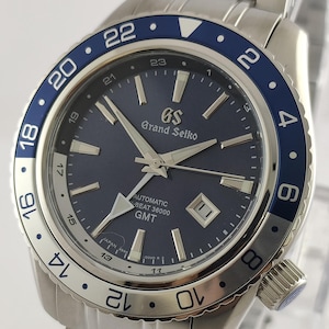 Puede incluir: Un reloj automático Grand Seiko de acero inoxidable con esfera azul marino y bisel azul y plateado. El reloj tiene una ventana de fecha y agujas luminosas. La correa del reloj es de acero inoxidable. La esfera muestra "Grand Seiko" y "AUTOMATIC BEAT 36000 GMT".