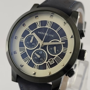 May include: A black Montblanc watch with a blue and cream face, featuring Roman numerals and subdials. The watch has a black bezel and a blue leather strap. The date is displayed at the 3 o'clock position.