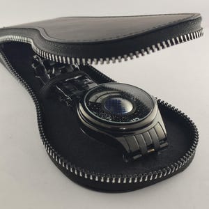 Puede incluir: Un reloj de pulsera negro con un dise&ntilde;o de esfera &uacute;nico, exhibido dentro de un estuche de cuero negro con cremallera plateada. El reloj tiene una correa oscura y una esfera redonda con un patr&oacute;n de estrellas. El estuche est&aacute; abierto, revelando el reloj.
