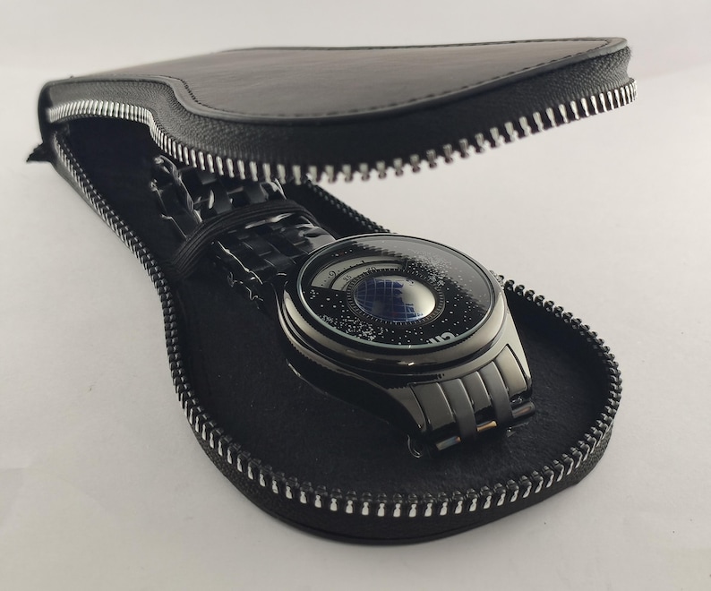 Puede incluir: Un reloj de pulsera negro con un dise&ntilde;o de esfera &uacute;nico, exhibido dentro de un estuche de cuero negro con cremallera plateada. El reloj tiene una correa oscura y una esfera redonda con un patr&oacute;n de estrellas. El estuche est&aacute; abierto, revelando el reloj.
