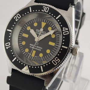 Può includere: Un orologio Seiko Submariner automatico nero e argento con lunetta e cinturino neri. Il quadrante dell'orologio è nero con indici e lancette gialle. Il testo "SEIKO AUTOMAT" e "SUBMARINER" è presente sul quadrante.