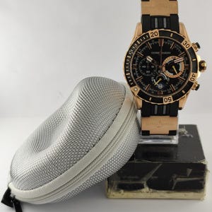 May include: A rose gold and black Ulysse Nardin chronograph watch with a black rubber strap, featuring a black dial with multiple sub-dials, a date window, and a rotating bezel, accompanied by a textured silver zippered carrying case.