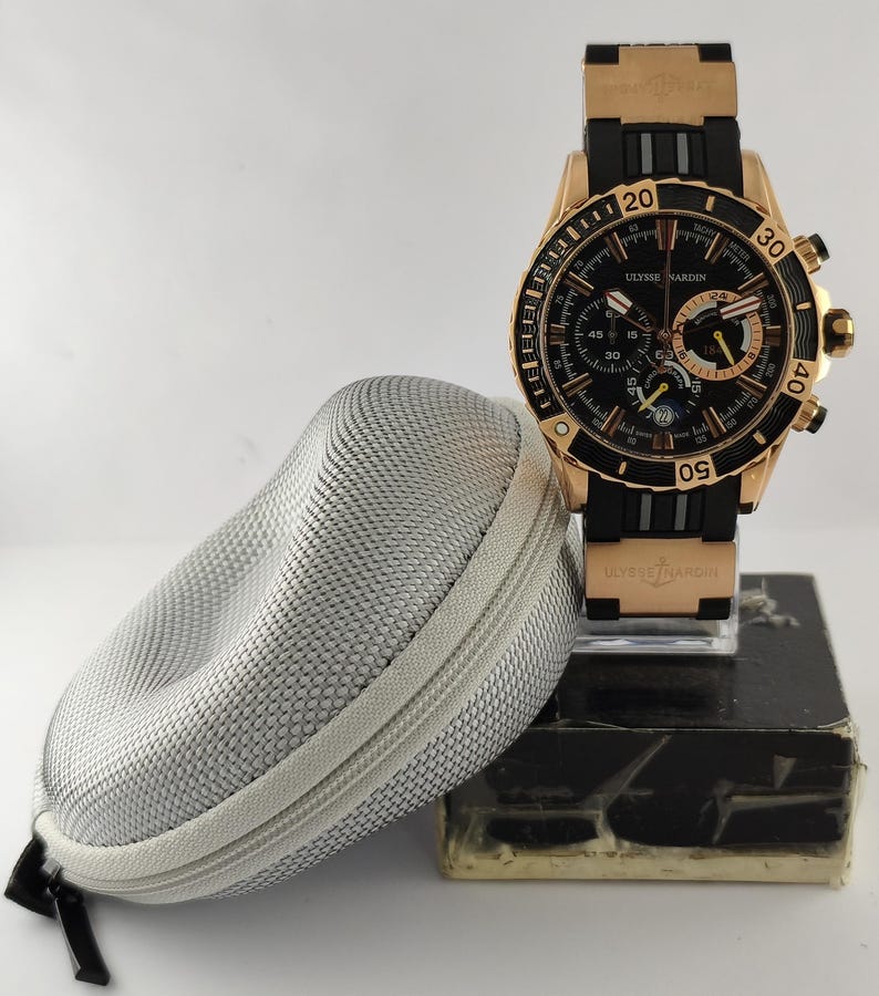 May include: A rose gold and black Ulysse Nardin chronograph watch with a black rubber strap, featuring a black dial with multiple sub-dials, a date window, and a rotating bezel, accompanied by a textured silver zippered carrying case.