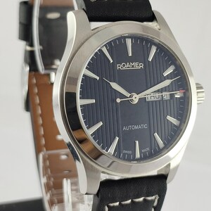 May include: A Roamer automatic wristwatch with a dark blue dial and silver hands. The watch features a black leather strap with white stitching. The watch face displays the day and date. The watch is Swiss made.